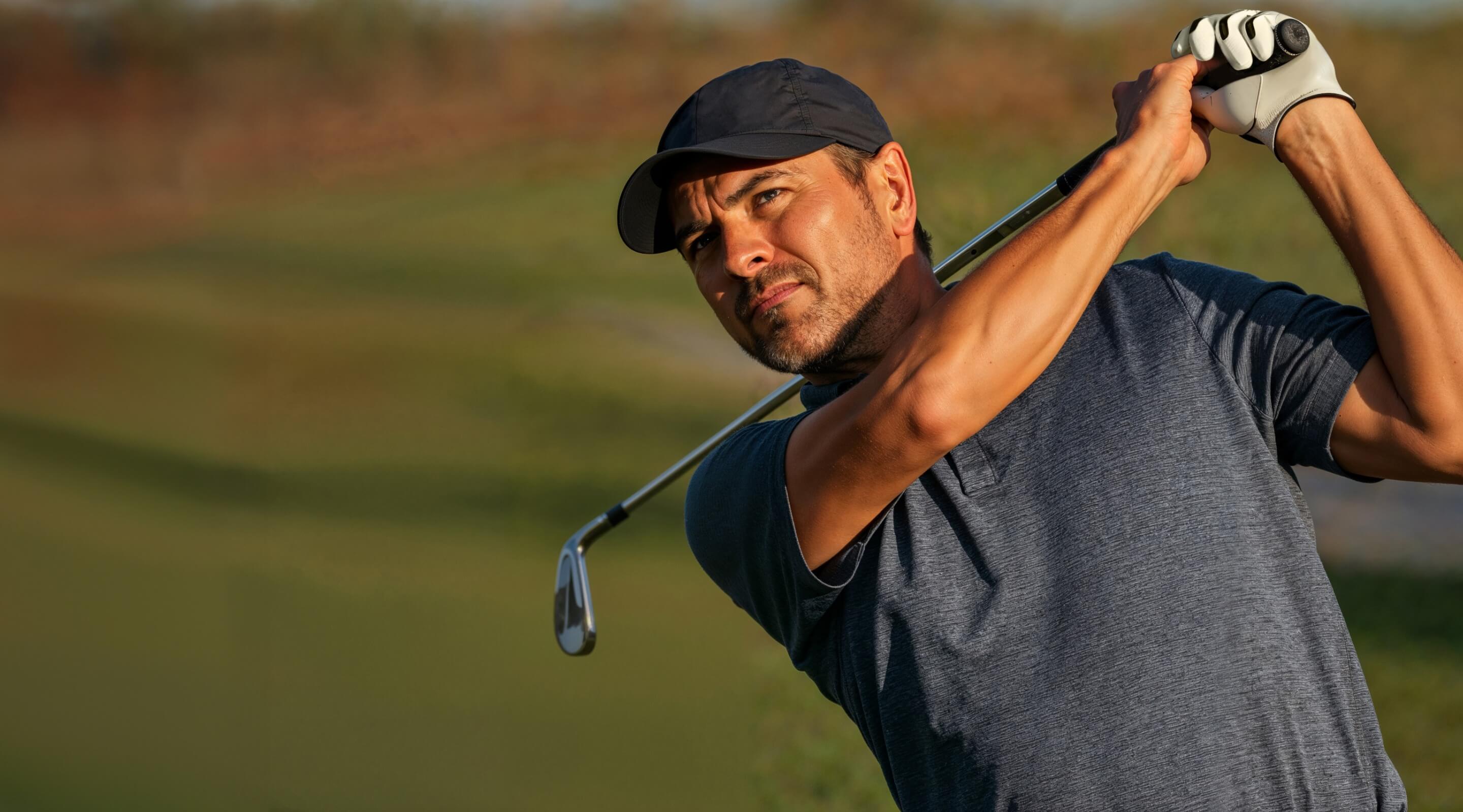 A man wearing a black T-shirt and black cap holds a golf club, preparing to take a swing on a green field.