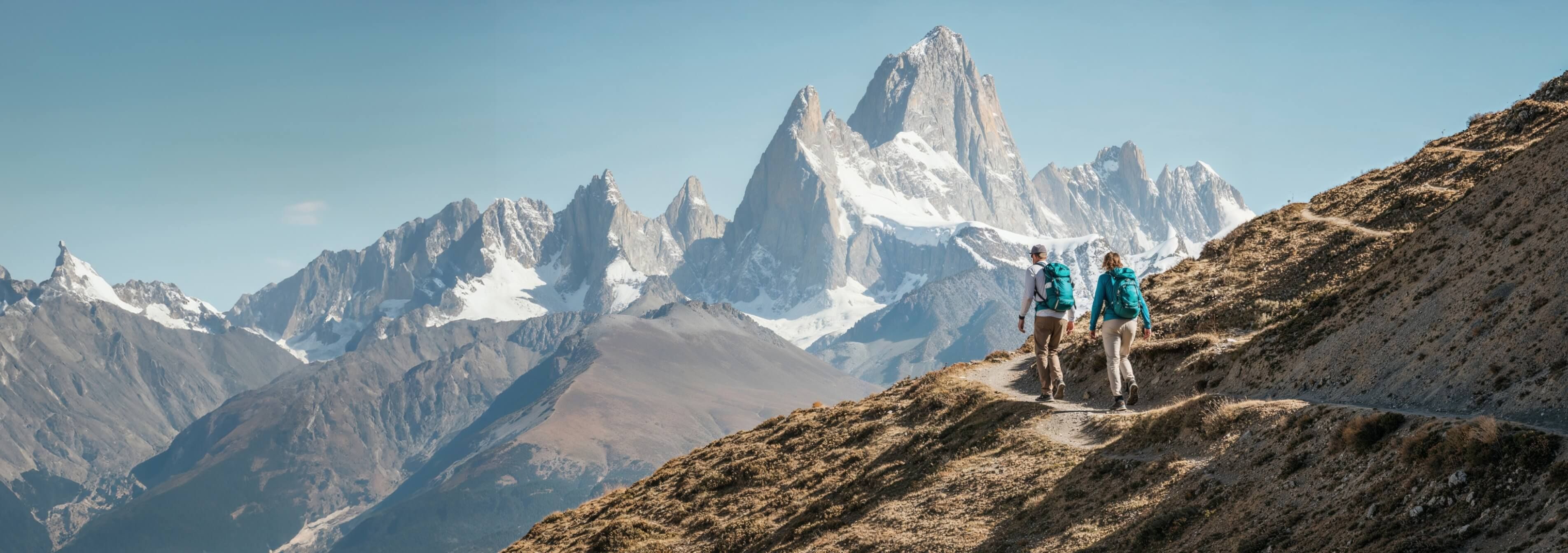Man and woman hiking along a rugged mountain trail, surrounded by steep cliffs and rocky terrain, with a vast view of distant peaks and a clear sky overhead.